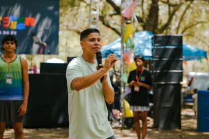 Person speaking at an outdoor event with attendees in the background.